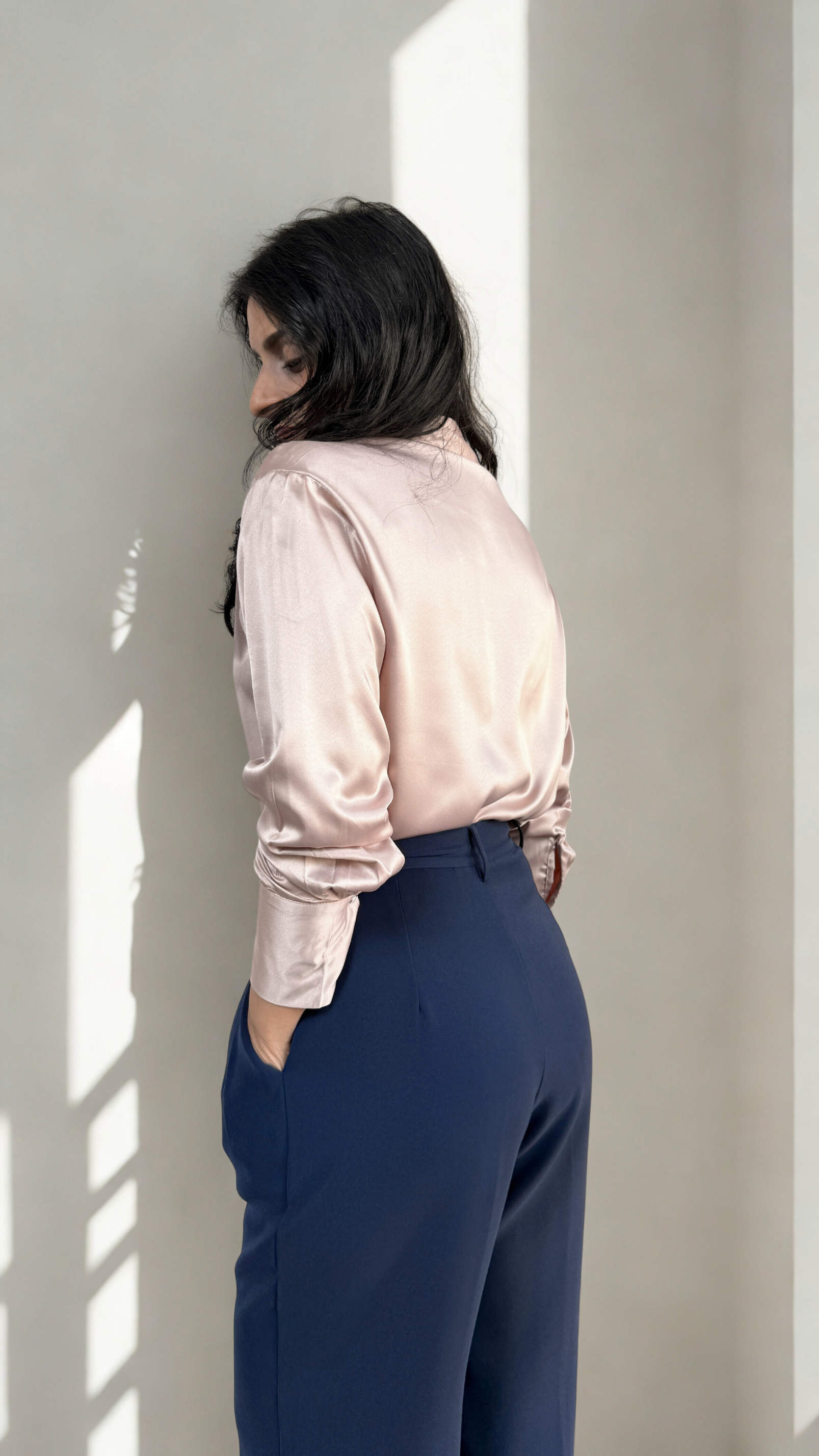 Person wearing a light pink blouse and blue pants standing against a neutral wall.